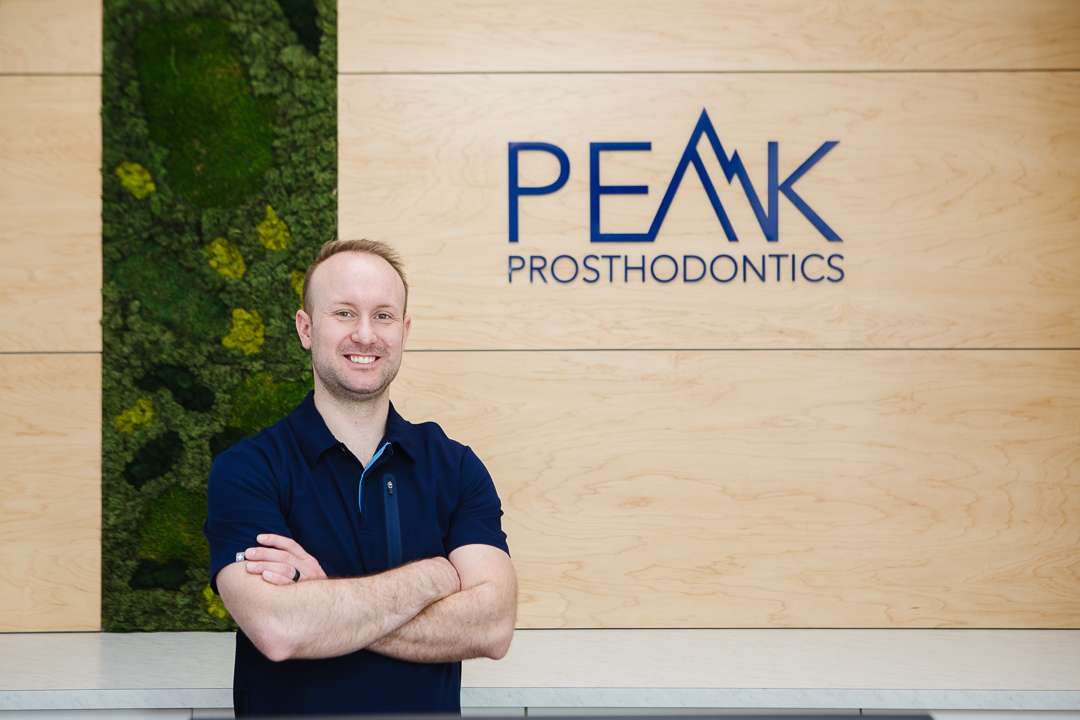 Male Dentist standing in front of decorative wall with Peak Prosthodontics sign on it
